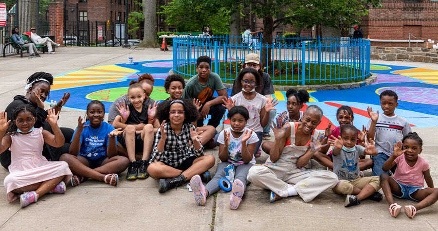 Volunteers and children sitting in front of a hand-painted mural on a sidewalk within a community park